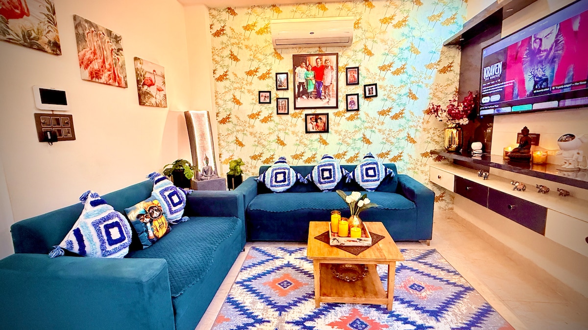 A shared living area features a blue sofa adorned with decorative cushions. A coffee table sits in the center, surrounded by patterned rugs. Artwork decorates the walls, and a smart TV is mounted on a cabinet, enhancing a vibrant yet cozy ambiance.