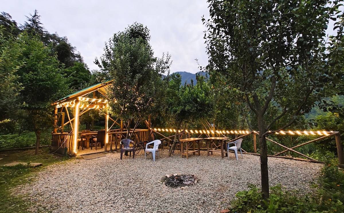 A cozy garden area features a gravel surface surrounded by lush greenery and trees. Seating arrangements include white chairs positioned around a central bonfire pit, while soft fairy lights are strung above, creating a serene ambiance in the outdoor space.