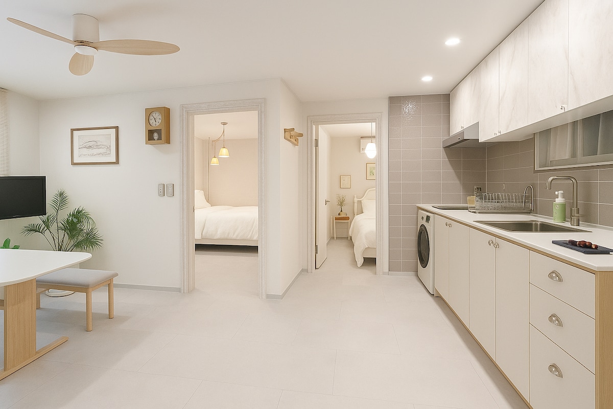 A modern kitchen features light cabinetry, a washing machine, and essential appliances, including a stove and sink. A spacious dining table sits nearby, and an open layout connects to inviting sleeping areas visible through adjacent doorways.