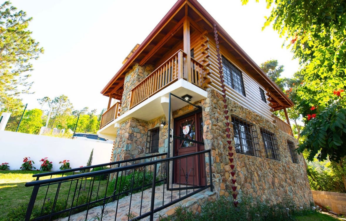 A charming villa is presented with a stone exterior and wooden balcony. The structure is surrounded by lush greenery, with a gently sloping pathway leading to the entrance. Large windows allow natural light to illuminate the interior spaces.
