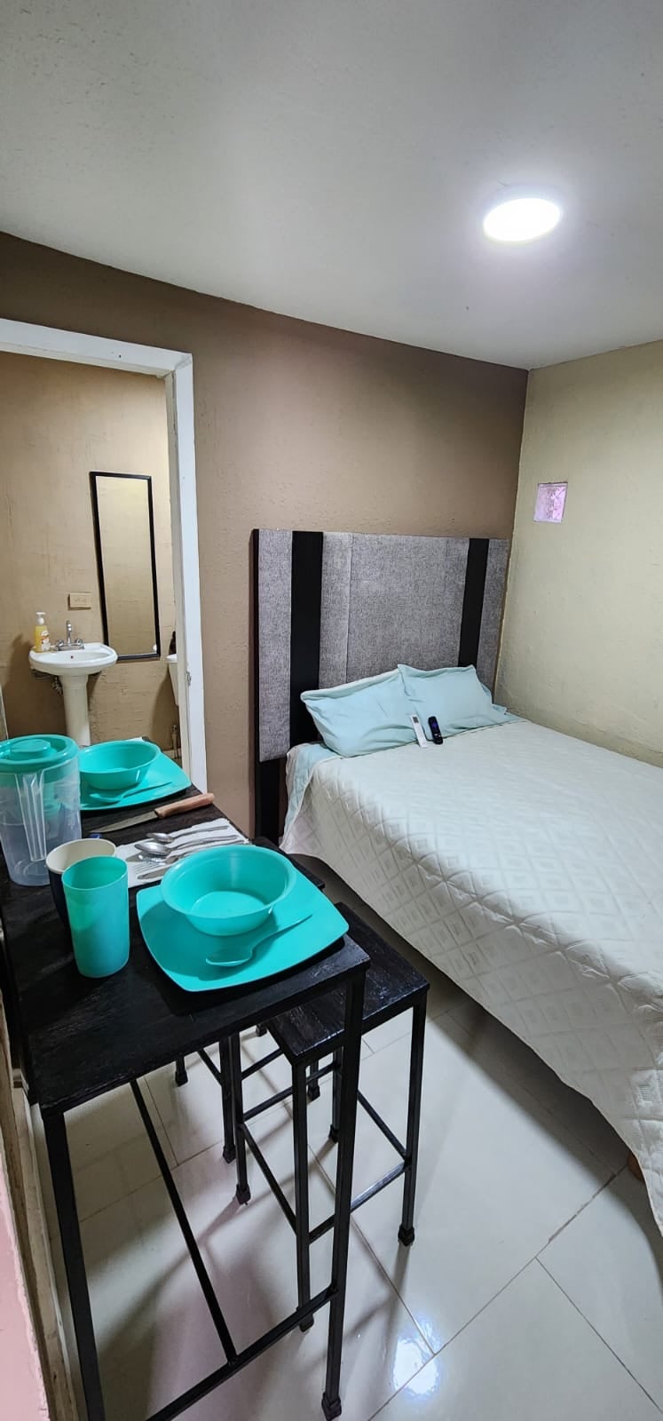 A compact studio space features a neatly arranged sleeping area with a light-colored quilted bedspread and two pillows. A small dining table with aqua-colored plates and glassware is positioned nearby, with a bright light fixture above illuminating the room.