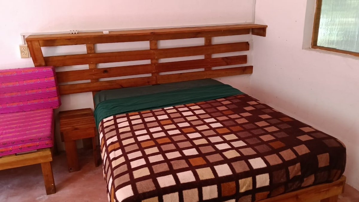 A spacious sleeping area is furnished with a large bed covered in a patterned blanket. A wooden headboard adds a rustic touch, and a small side table is positioned beside the bed. A pink seat with striped fabric is available, and a window provides natural light.