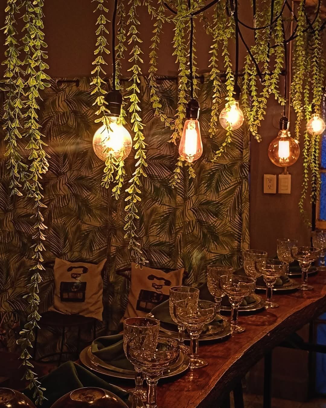 A dining area is showcased with a wooden table set for a meal, featuring glassware and plates. Hanging lights illuminate the space, surrounded by flowing greenery. The backdrop displays a patterned curtain with leafy designs, adding a natural element to the atmosphere.