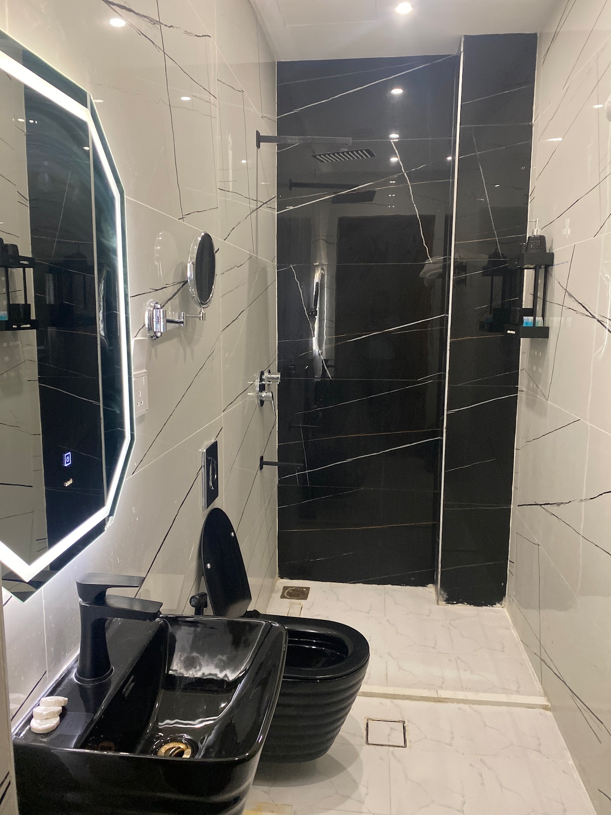 A modern bathroom is presented with sleek black and white marble finishes. An elegant sink and toilet are positioned against the wall. Stylish lighting incorporates a mirror, while a spacious shower area is enclosed by glass panels.