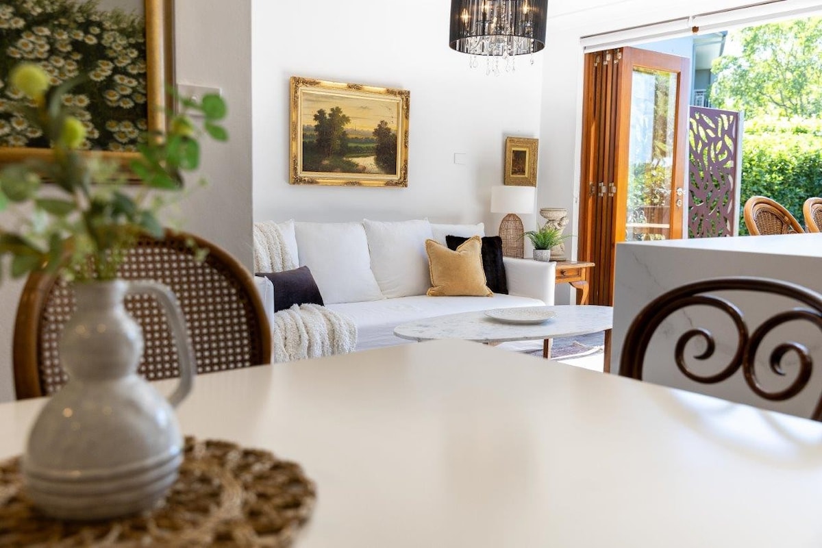 A comfortable living space features a light-colored sofa adorned with soft cushions. A vintage painting hangs above, while bi-fold doors open to reveal lush greenery outside. A dining table with a woven placemat and a vase adds to the inviting atmosphere.