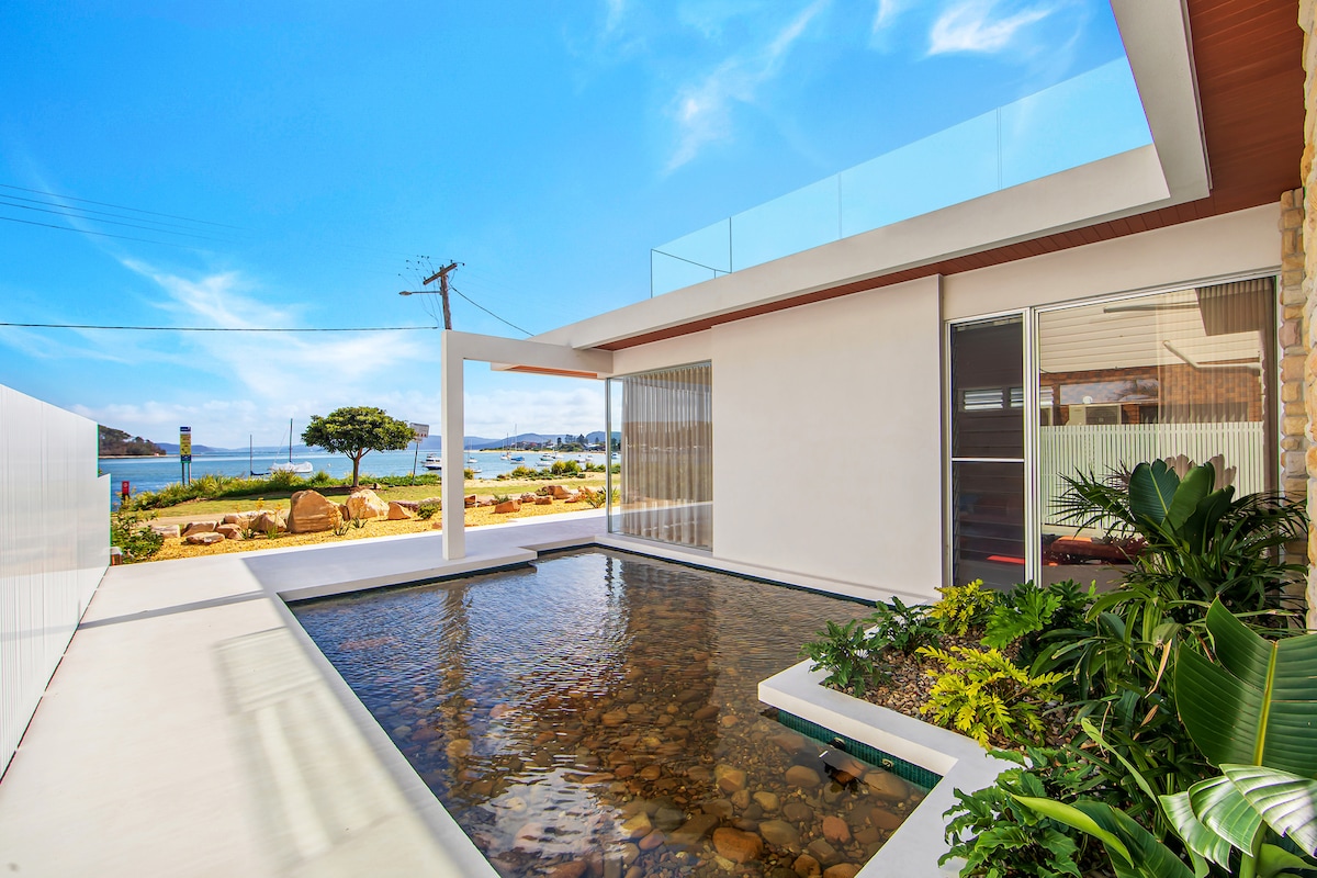 A serene outdoor area features a water feature surrounded by lush greenery, providing a calming atmosphere. Floor-to-ceiling glass panels enhance the views of the beach and surrounding landscape, while the bright blue sky adds a touch of openness to the space.