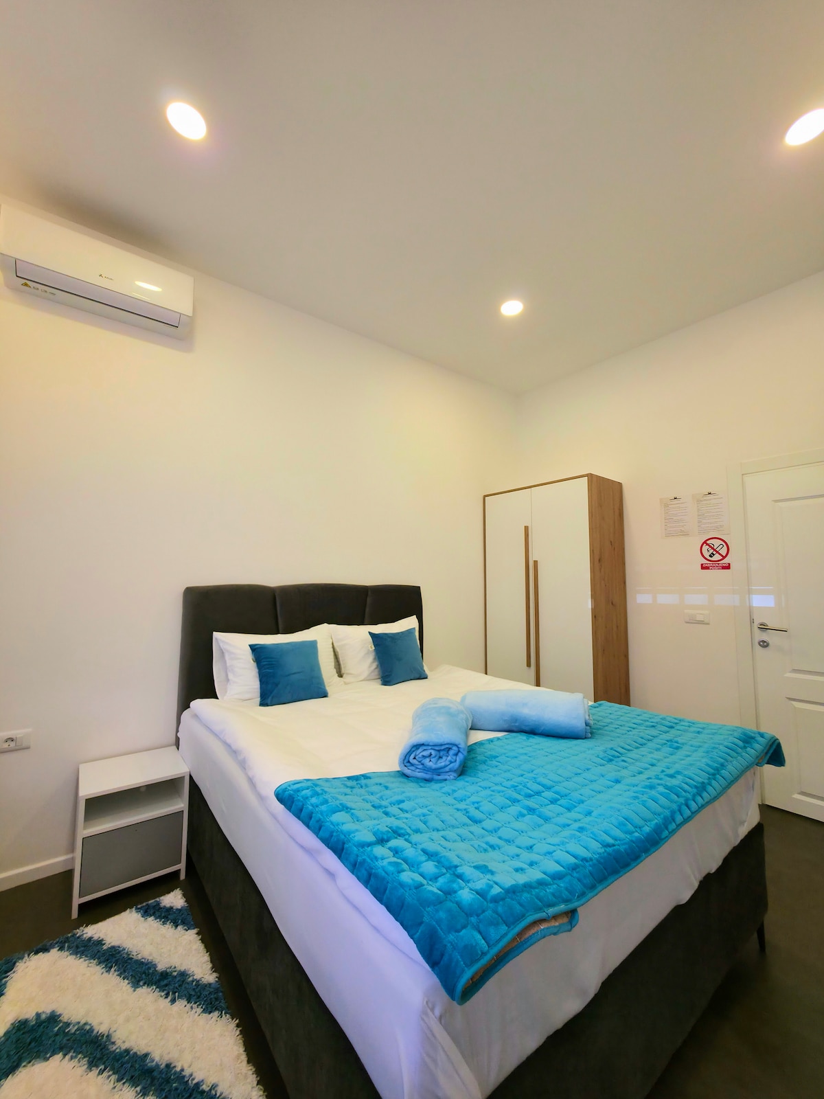 A modern bedroom features a spacious bed with white linens and decorative blue accents. A textured blanket is neatly draped at the foot of the bed. Adjacent, a wardrobe and a nightstand are visible. Light from overhead fixtures creates a bright and airy atmosphere.