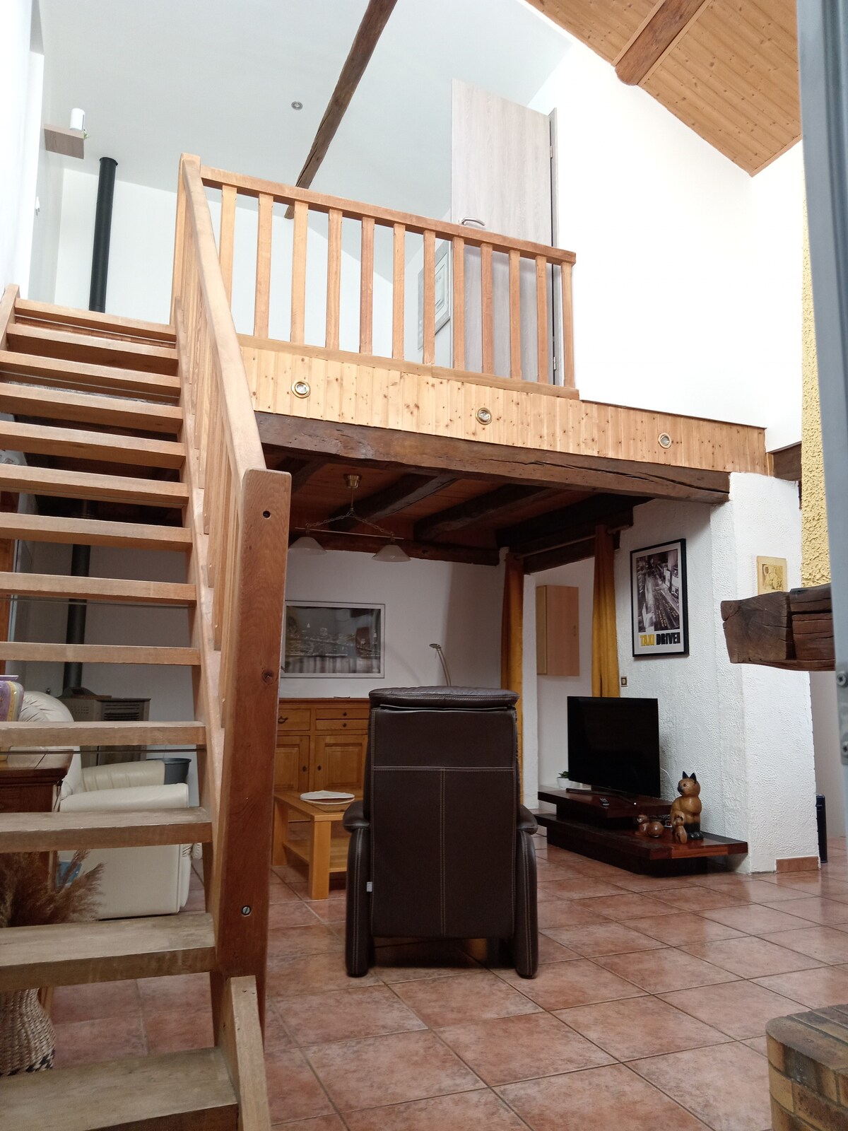 The interior features an open layout with wooden beams and a staircase leading to an upper level. A cozy sitting area is visible, equipped with a television and wooden furniture. Natural light floods the space, highlighting the warm tones of the tiles and furnishings.