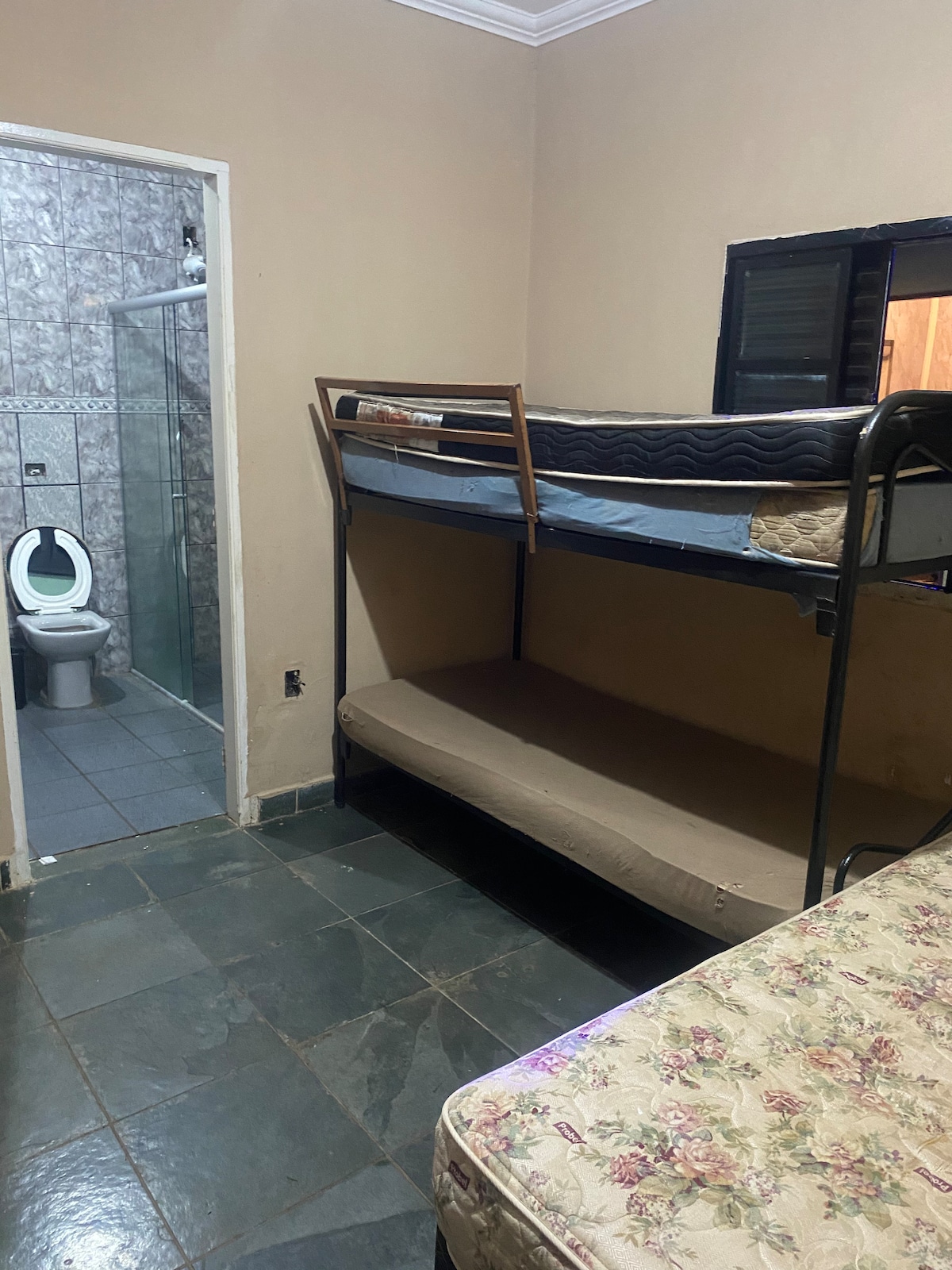 A compact bedroom contains a bunk bed with a mattress on the top and a mattress on a lower frame. A double bed with floral bedding occupies the adjacent space. A bathroom entrance is visible, leading to tiled flooring and walls.