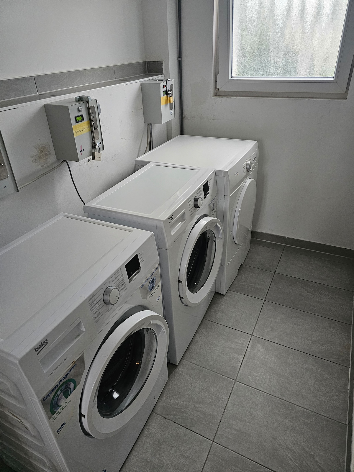 The laundry area features three white appliances: two washing machines and one dryer, positioned against a clean, tiled wall. A window allows natural light to enter, illuminating the space. Each machine has a simple control panel for user convenience.