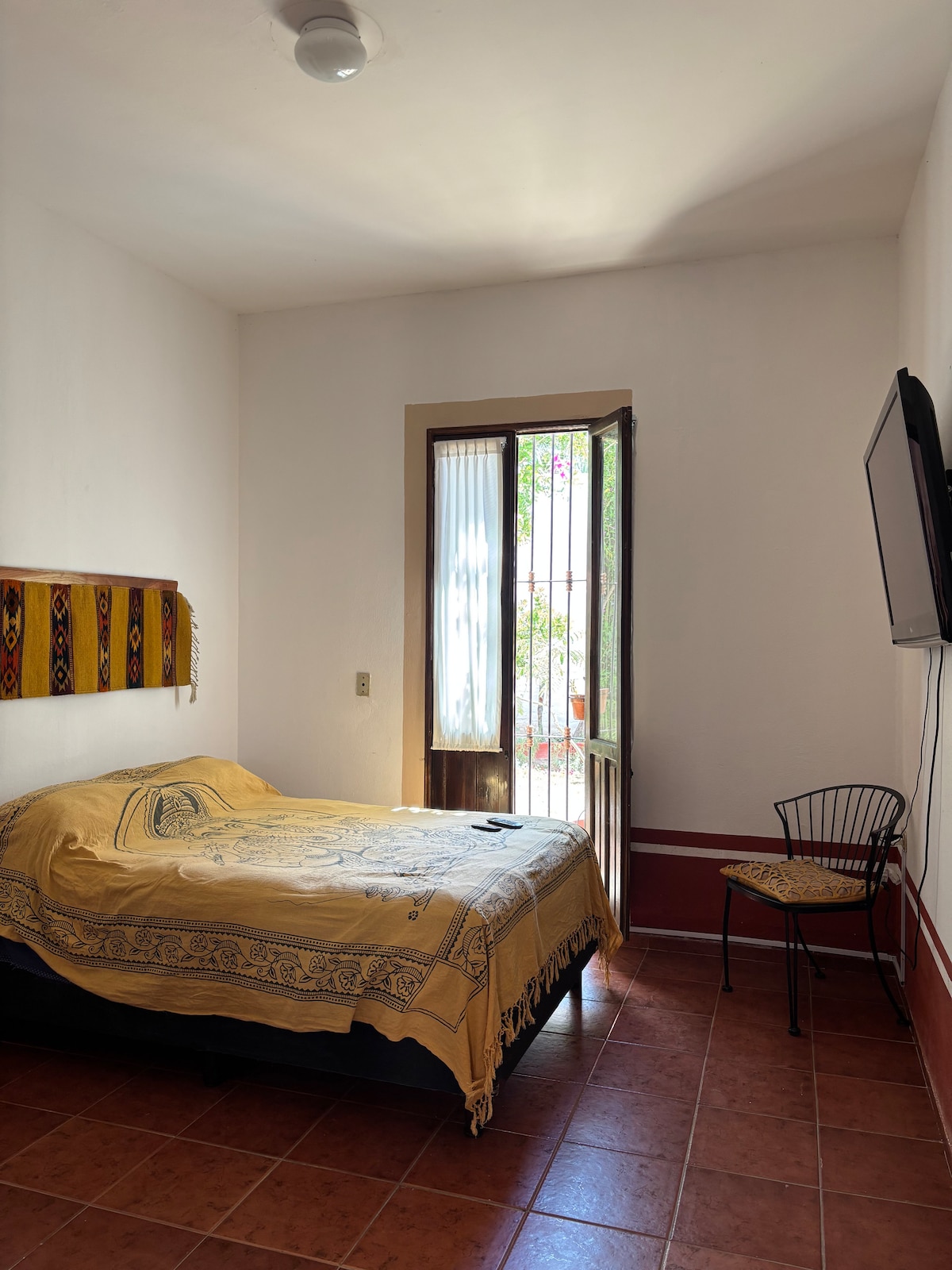A cozy bedroom is presented featuring a spacious bed adorned with a decorative golden blanket. Natural light enters through the window beside a simple black chair. A wall-mounted television adds convenience, while the tiled floor complements the warm ambiance.