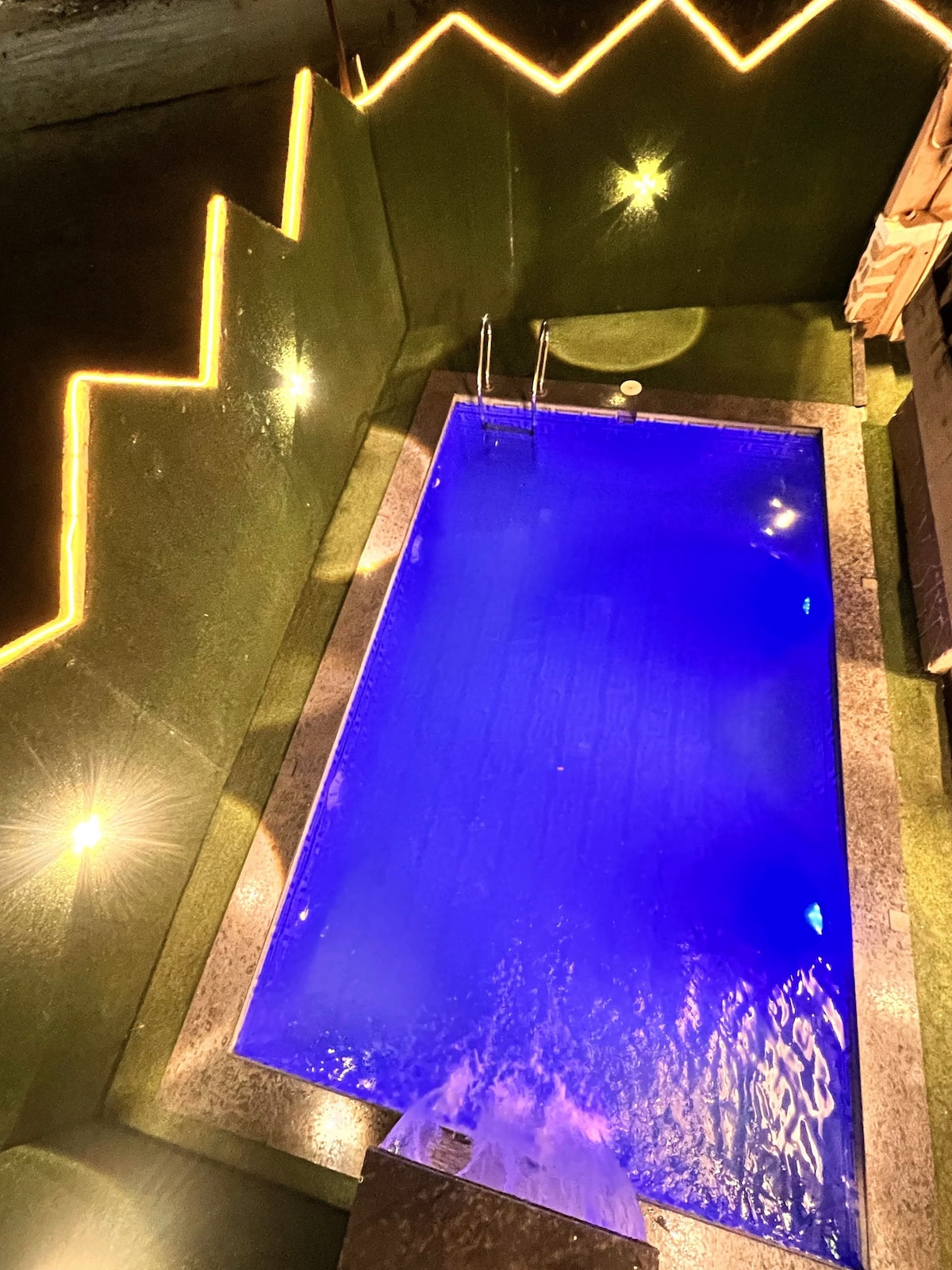 An illuminated swimming pool features shimmering blue water, surrounded by greenery and stylish lighting. The pool's edges are lined with smooth stone, enhancing the modern design. Soft ambient lights contribute to a tranquil nighttime atmosphere.