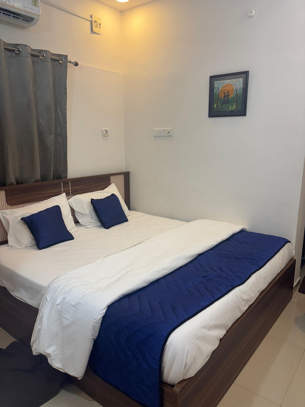 A cozy sleeping area is presented, featuring a spacious bed dressed in a white comforter with a navy blue accent blanket. Two pillows adorned in navy blue rest against the headboard. A small piece of artwork depicting nature is hung on the wall, adding a subtle decorative touch.