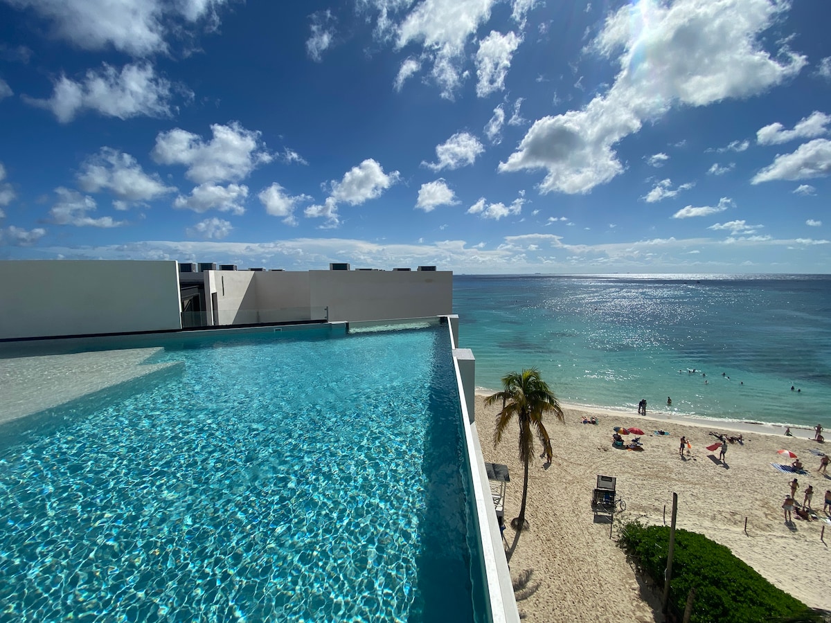 An expansive infinity pool is visible, merging seamlessly with the horizon where the ocean meets the sky. White sand beach is seen below, dotted with beachgoers. Bright blue water reflects the clouds above, creating a serene scene.