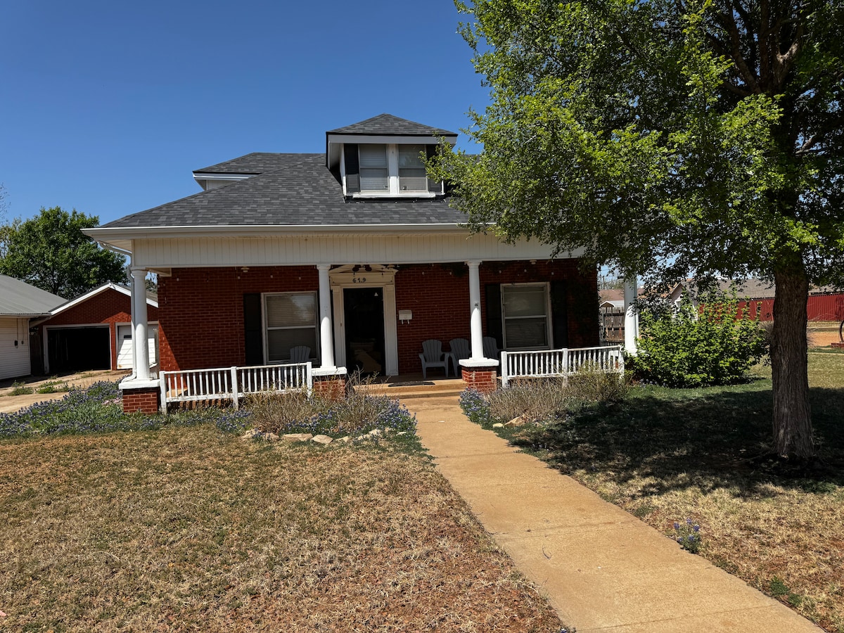 A charming two-story home features a covered front porch with seating, surrounded by green grass and landscaping. A concrete pathway leads to the entrance, bordered by mulch and flowering plants. A large tree provides shade, enhancing the outdoor appeal of the property.