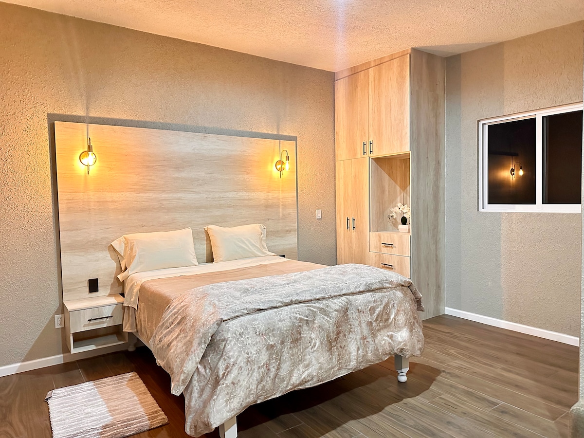 A cozy sleeping area features a plush bed with neutral bedding, complemented by softly glowing pendant lights on either side. A nightstand is positioned next to the bed, while a spacious wardrobe is built into the wall. A window allows natural light to fill the room.