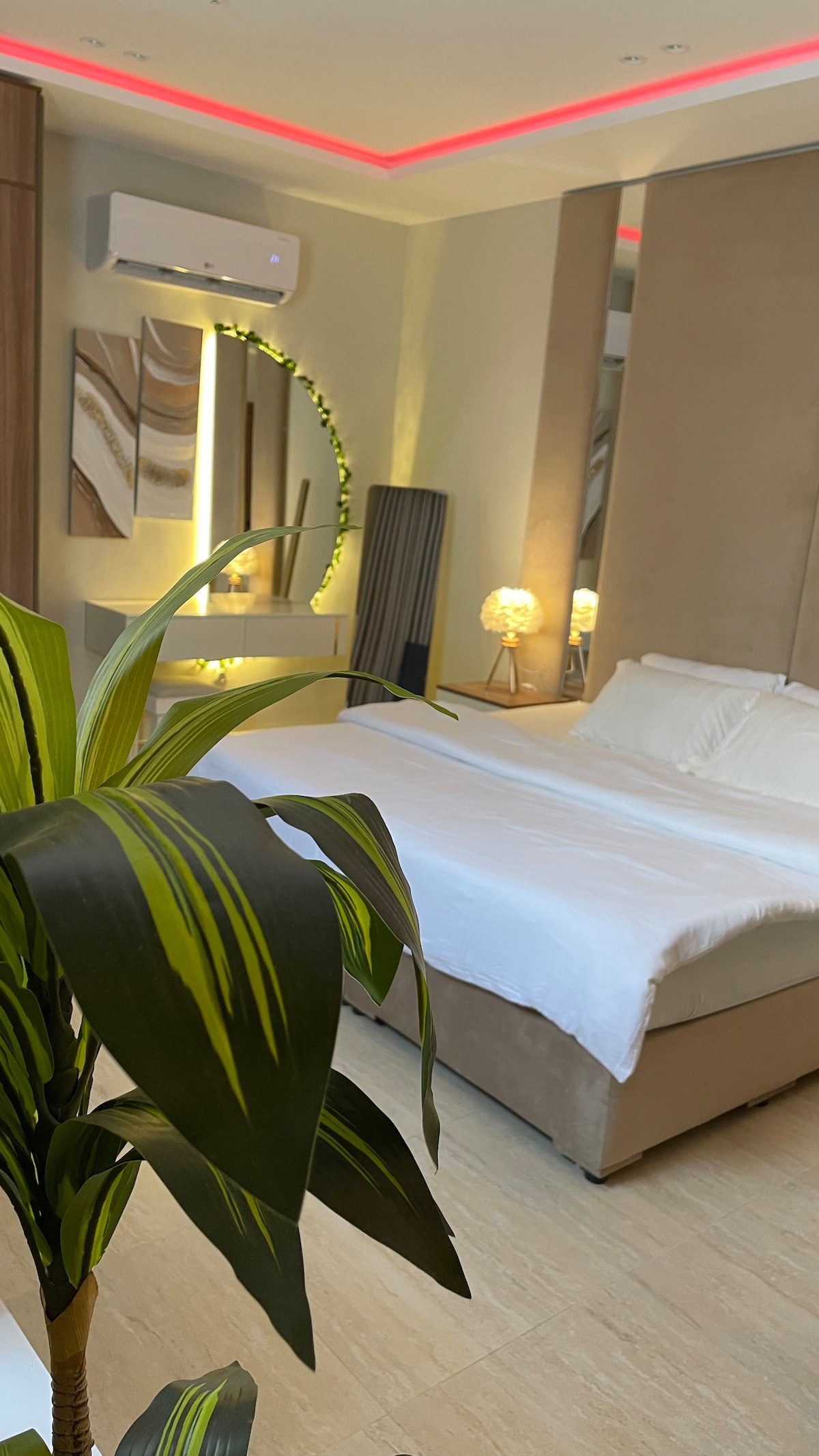A spacious sleeping area is highlighted by a large bed with a light-colored duvet. Decorative lighting and modern artwork illuminate the room. A potted plant adds a touch of greenery, contributing to the overall serene atmosphere.