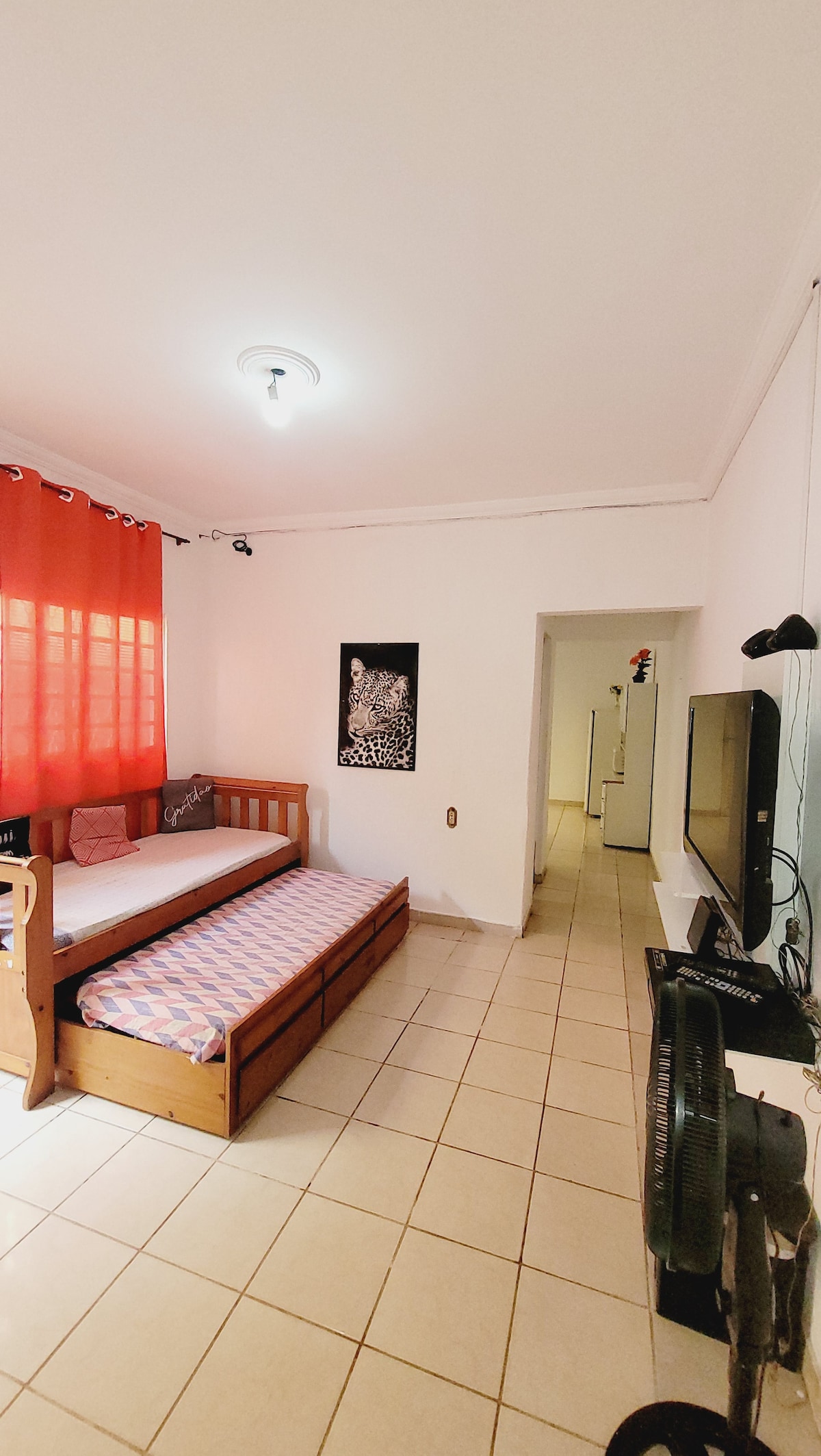 A well-lit bedroom features a wall-mounted television and a comfortable daybed with an additional trundle positioned beside it. Bright red curtains filter natural light from the window. The room's tiled floor and white walls enhance its spacious feel, and a standing fan is placed nearby.