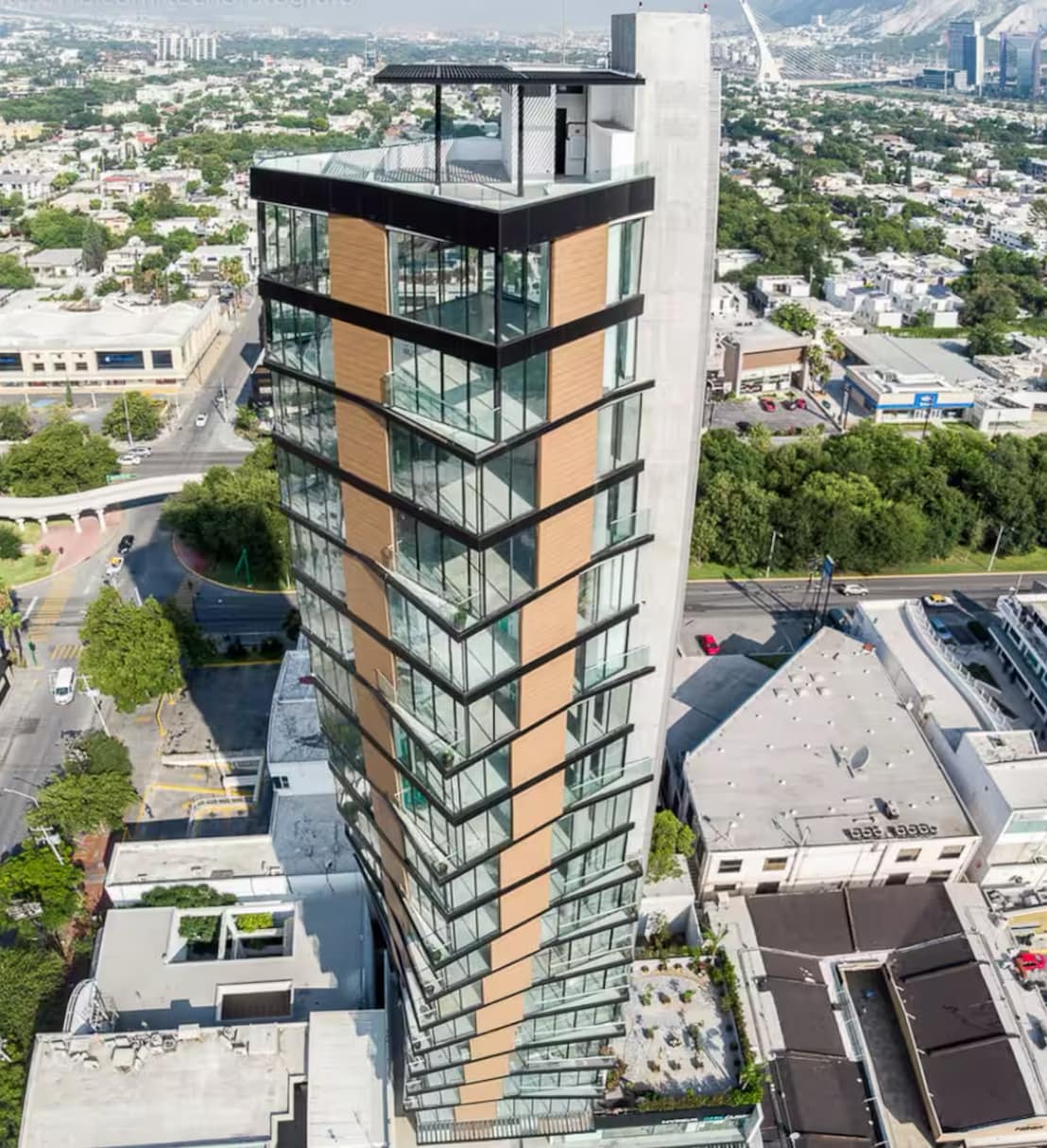 An aerial view of a modern, multi-story building with a striking architectural design. The exterior features a combination of glass and textured materials, while surrounding greenery and urban landscape provide context. The rooftop offers a spacious terrace area, visible at the top of the structure.