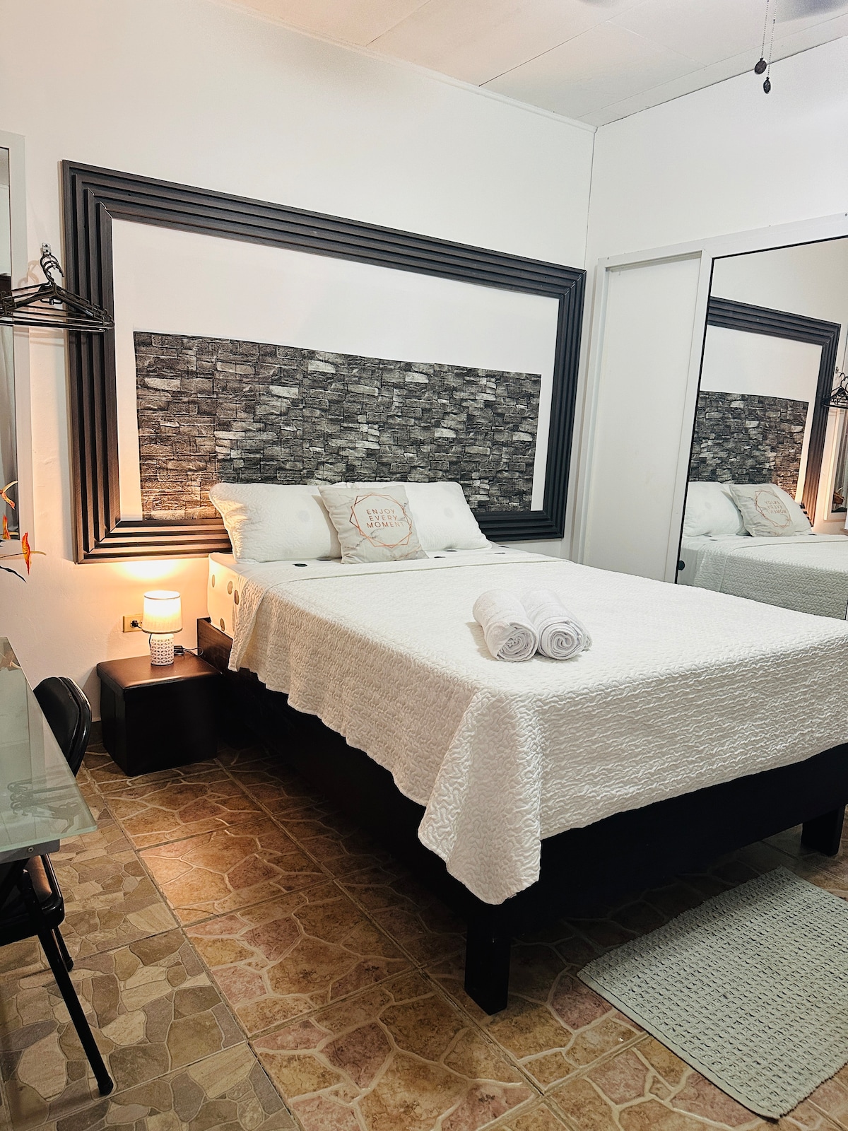 A well-appointed sleeping area features a plush bed covered with a textured white blanket. A decorative wall serves as a backdrop, while mirrored closets enhance the sense of space. A small nightstand holds a lamp, contributing to the room's functionality.