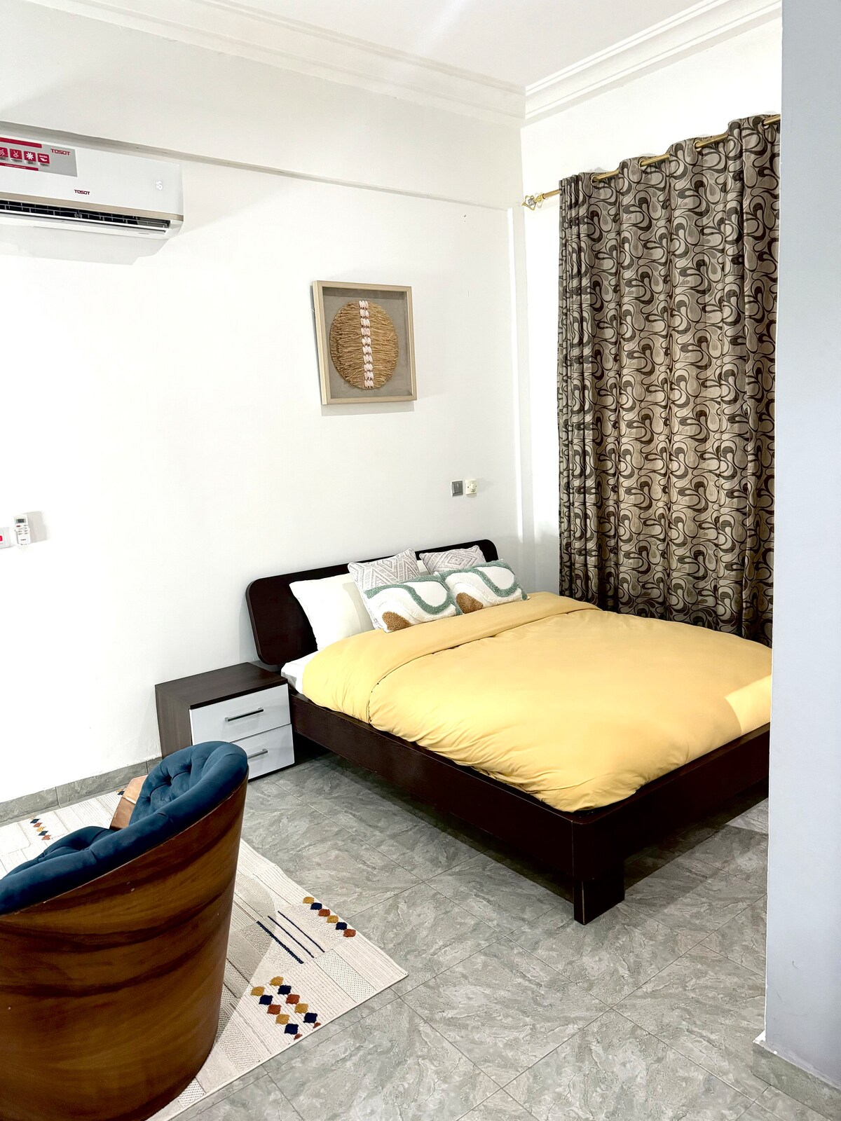 A cozy sleeping area is presented with a spacious bed featuring a yellow comforter. A patterned curtain hangs beside the bed, allowing soft light to enter. A wooden chair is positioned near a bedside table, adding to the room's functionality and comfort.