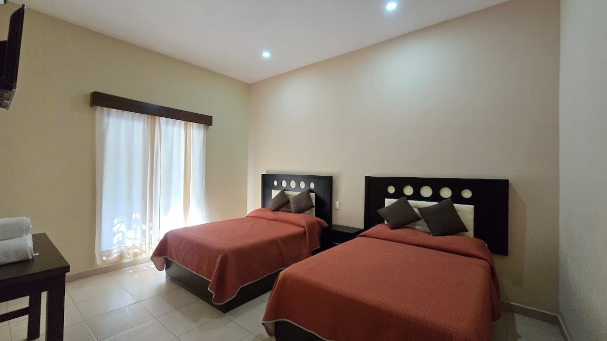 A brightly lit room features two spacious beds with orange bedspreads, accented by decorative pillows. Natural light enters through a sheer-curtained window, while a small table is positioned beside the beds. A television is mounted on the wall opposite the beds.