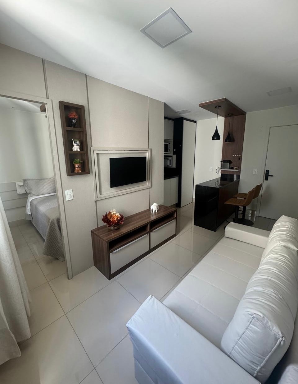 A well-organized living space features a neutral palette with glossy tiled floors. A small couch sits opposite a wall-mounted television, and a kitchenette with modern appliances is visible in the background. Decorative elements, such as a small vase, add a touch of warmth.