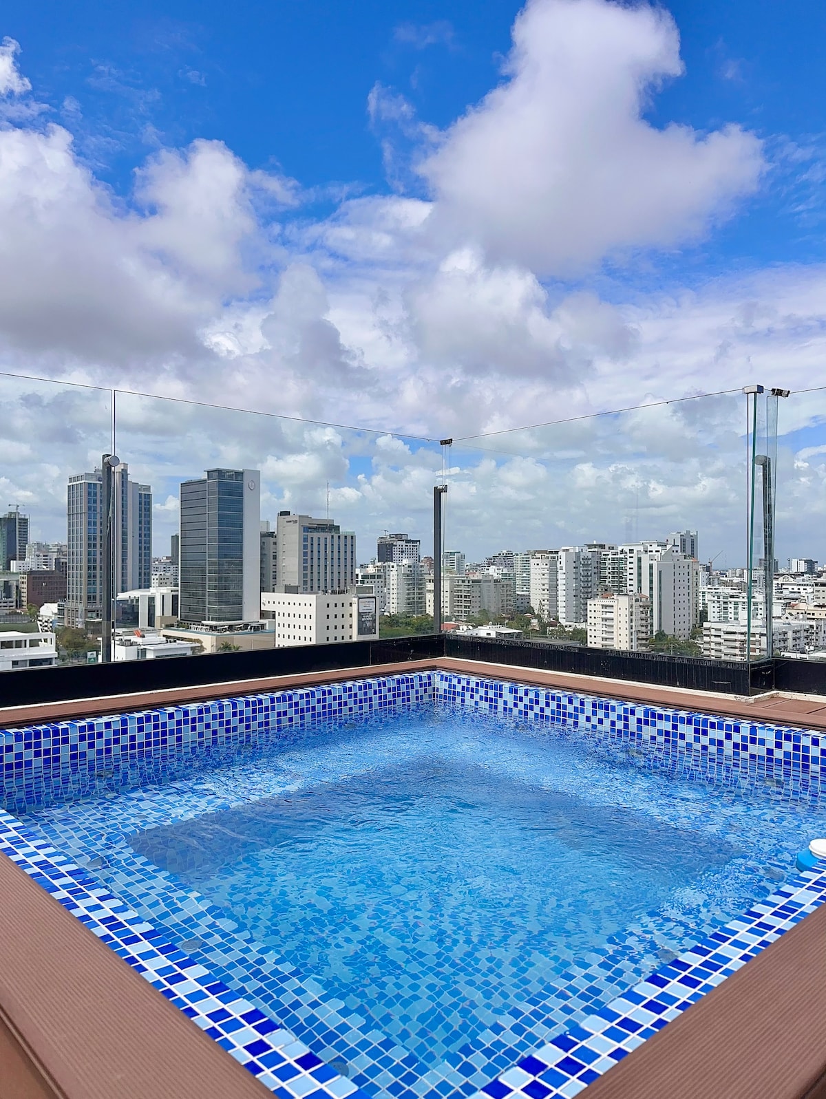A rooftop pool is showcased against a backdrop of the city skyline, featuring sparkling blue water surrounded by vibrant blue-tiled accents. The expansive view reveals a mix of modern buildings under a partly cloudy sky.