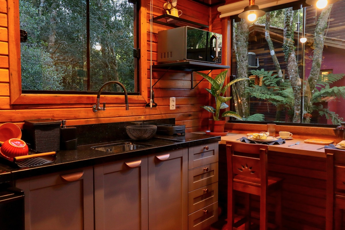 A functional kitchen space is equipped with a sink, microwave, and ample countertop area. Wooden cabinetry complements the natural surroundings visible through a large window, allowing for an outdoor view. A small dining table and chairs are positioned for enjoying meals.