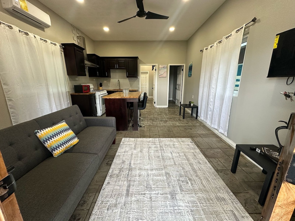 A spacious living area is presented with a gray couch featuring a colorful throw pillow. Light-colored walls and large windows allow natural light to fill the room. A dining area with a bar counter and modern kitchen cabinets is visible in the background, complementing the overall layout.
