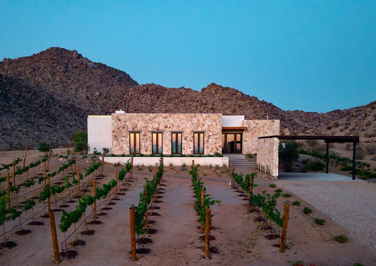 The villa features a modern stone exterior, harmoniously integrated with the desert landscape. A vineyard with rows of grapevines lies in the foreground, with rugged mountains providing a scenic backdrop at dusk. Warm light glows from the windows, inviting relaxation.