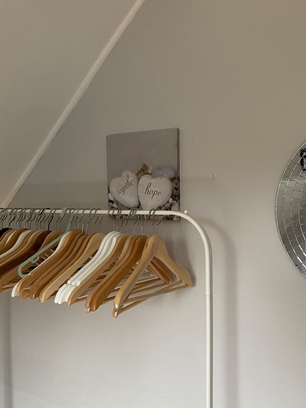A clothing rack displays several wooden hangers, showcasing a neat and organized space. Above the rack, a decorative wall art piece is presented, featuring two hearts with the words 'together' and 'hope.' A round mirror reflects light, adding to the overall brightness of the room.