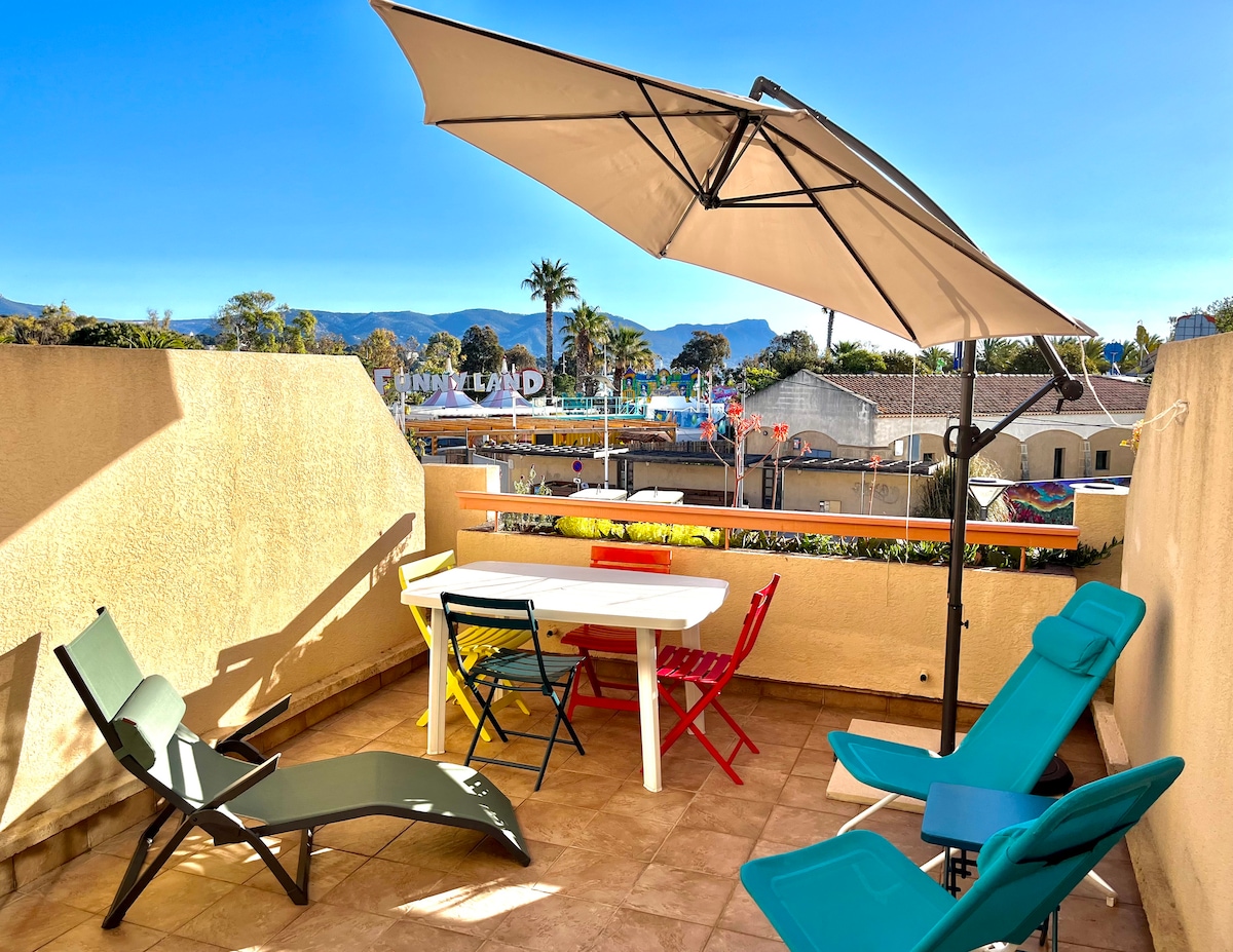 A sunlit terrace features a white table surrounded by colorful chairs and lounging chairs. An umbrella provides shade, enhancing the outdoor space. Scenic views of palm trees and distant mountains are visible, framed by a clear blue sky.