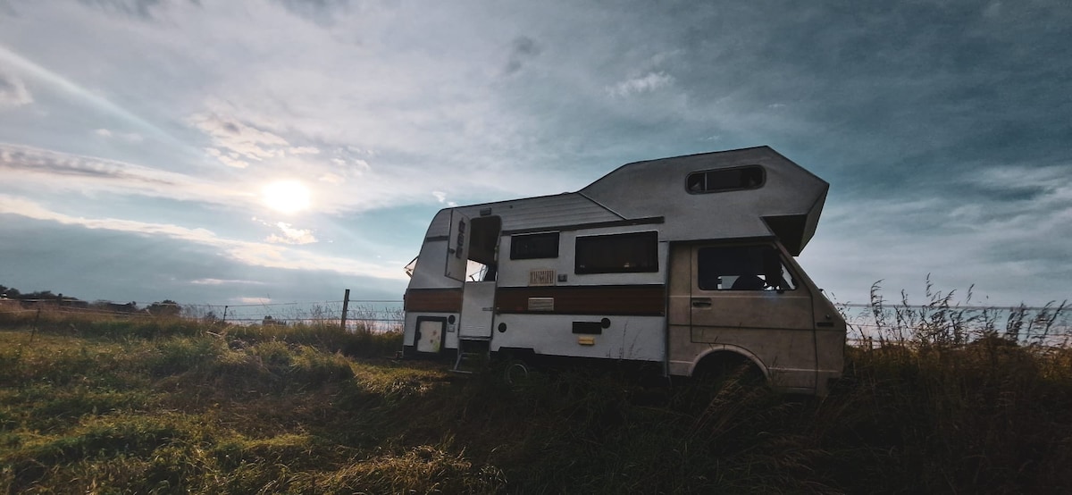 A vintage camper van is situated in a grassy field, surrounded by open space. The vehicle features distinct exterior colors and a unique shape, with windows reflecting the soft light of an overcast sky. The sun hangs low, casting a serene ambiance.
