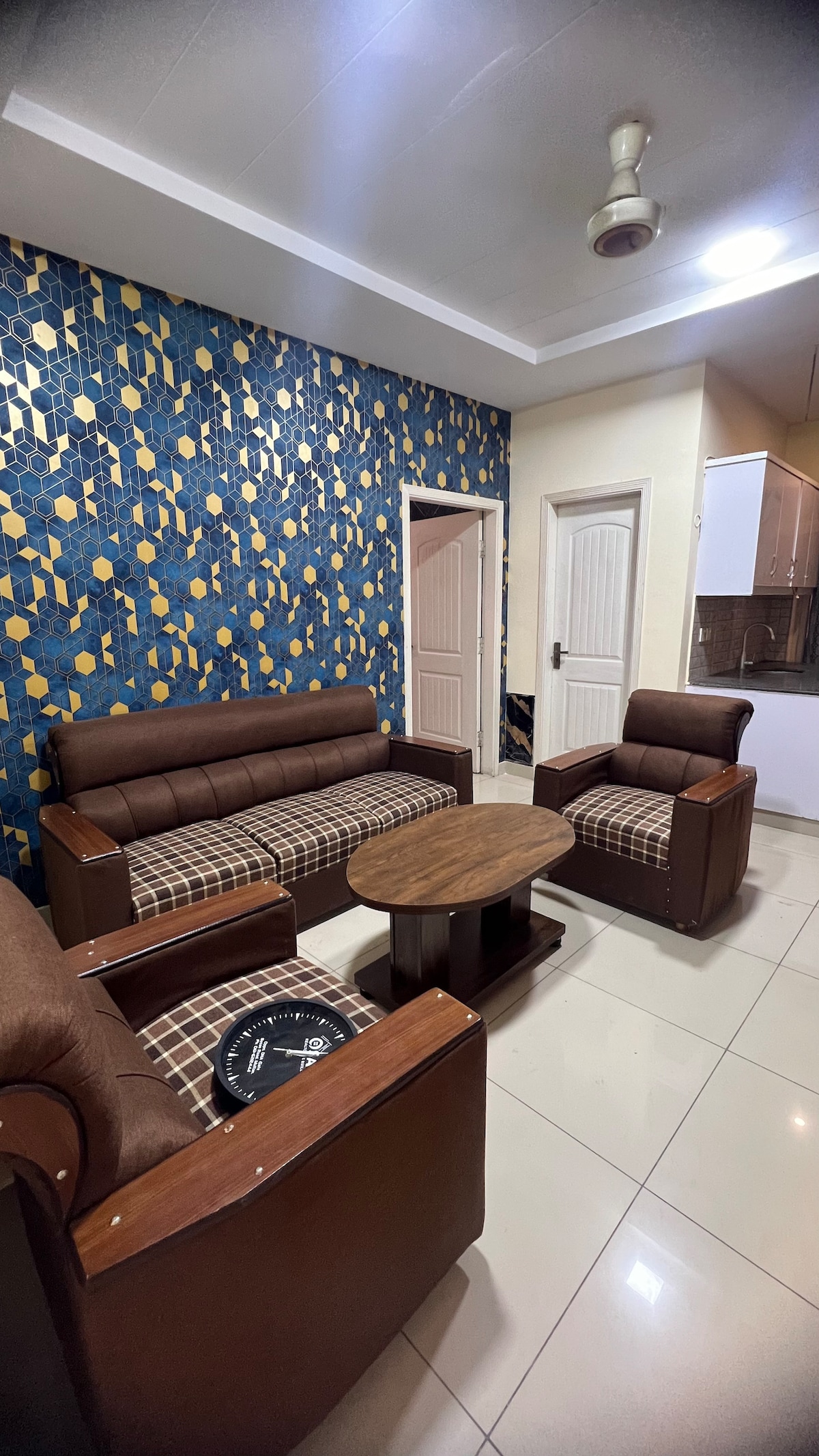 A modern living area is featured with a patterned accent wall in blue and gold tones. Brown leather sofas and armchairs surround a wooden coffee table. Light tiles reflect the space's brightness, complemented by clear doorways leading to additional areas.