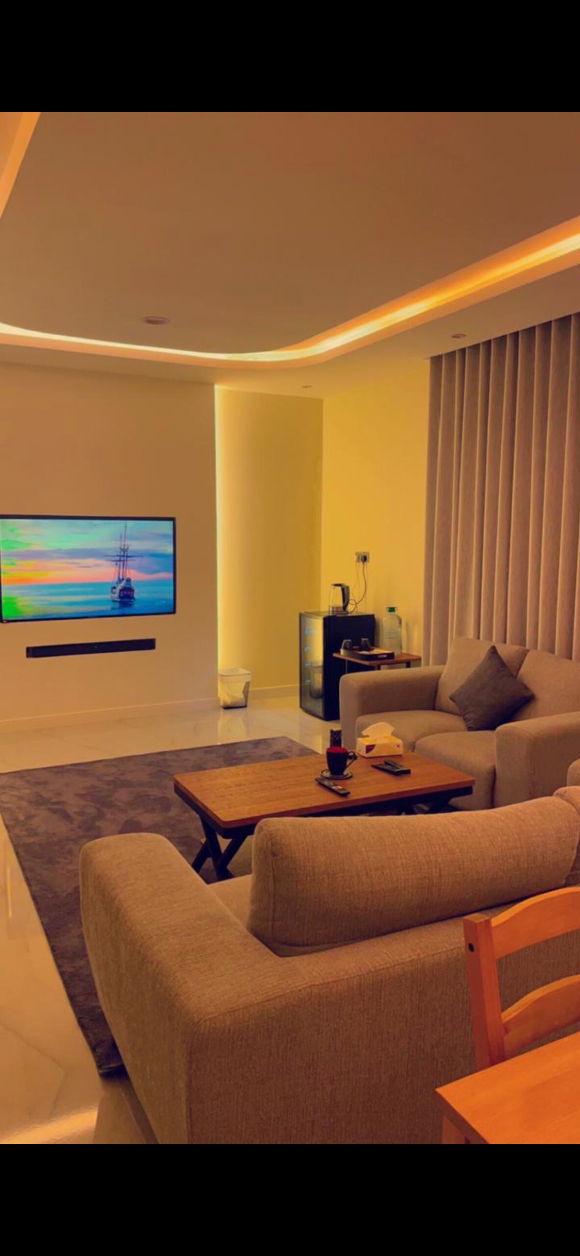 A modern living area features a large smart television mounted on the wall, displaying a colorful seaside scene. Soft, neutral-colored sofas surround a small coffee table, enhancing the space. Tall curtains frame the window, adding to the overall serene ambiance.