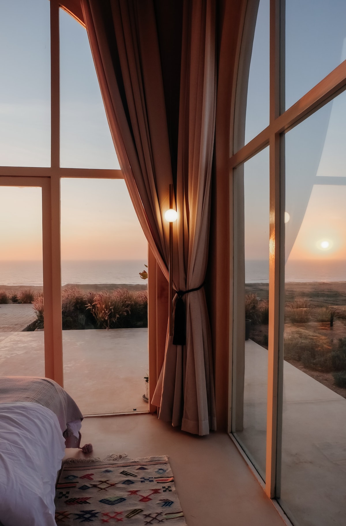 A serene bedroom is framed by large glass windows, offering a view of the sunset reflecting on the sea. Soft curtains partially drape over the glass, creating a gentle contrast with the bright colors of twilight. A cozy bed with neutral bedding is paired with a patterned rug beneath.