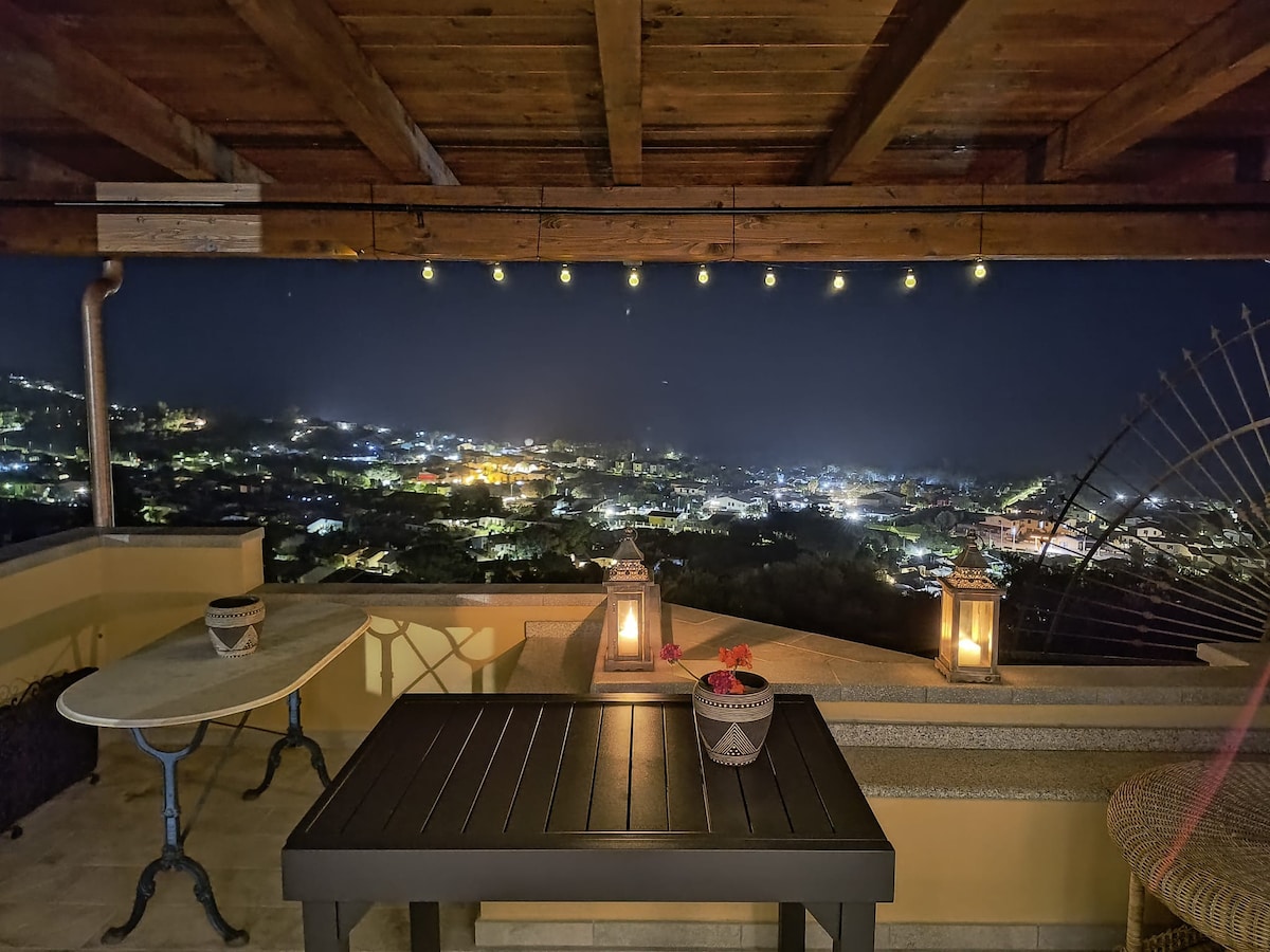 An outdoor terrace is illuminated by soft string lights, featuring a dark table and two decorative lanterns. The view showcases a city filled with twinkling lights, creating a peaceful night ambiance. The wooden beams overhead enhance the intimate atmosphere.
