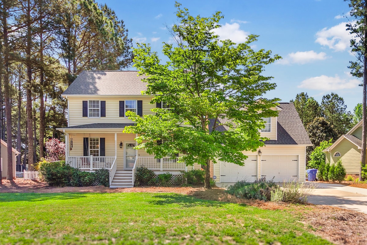 The exterior of a two-story house is showcased, featuring a welcoming front porch with white railings. A large tree provides shade in the front yard, complemented by green grass and a gravel driveway leading to a two-car garage.