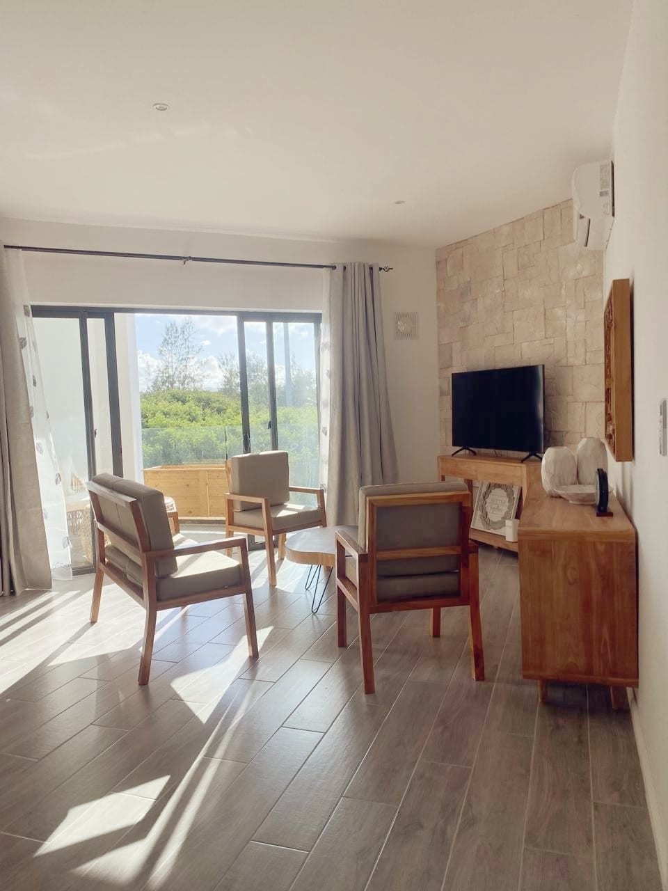 A bright living space is highlighted by large glass sliding doors, allowing natural light to fill the room. Two armchairs and a wooden coffee table are positioned thoughtfully, and a mounted television is visible on a textured stone wall, offering a comfortable area to relax.