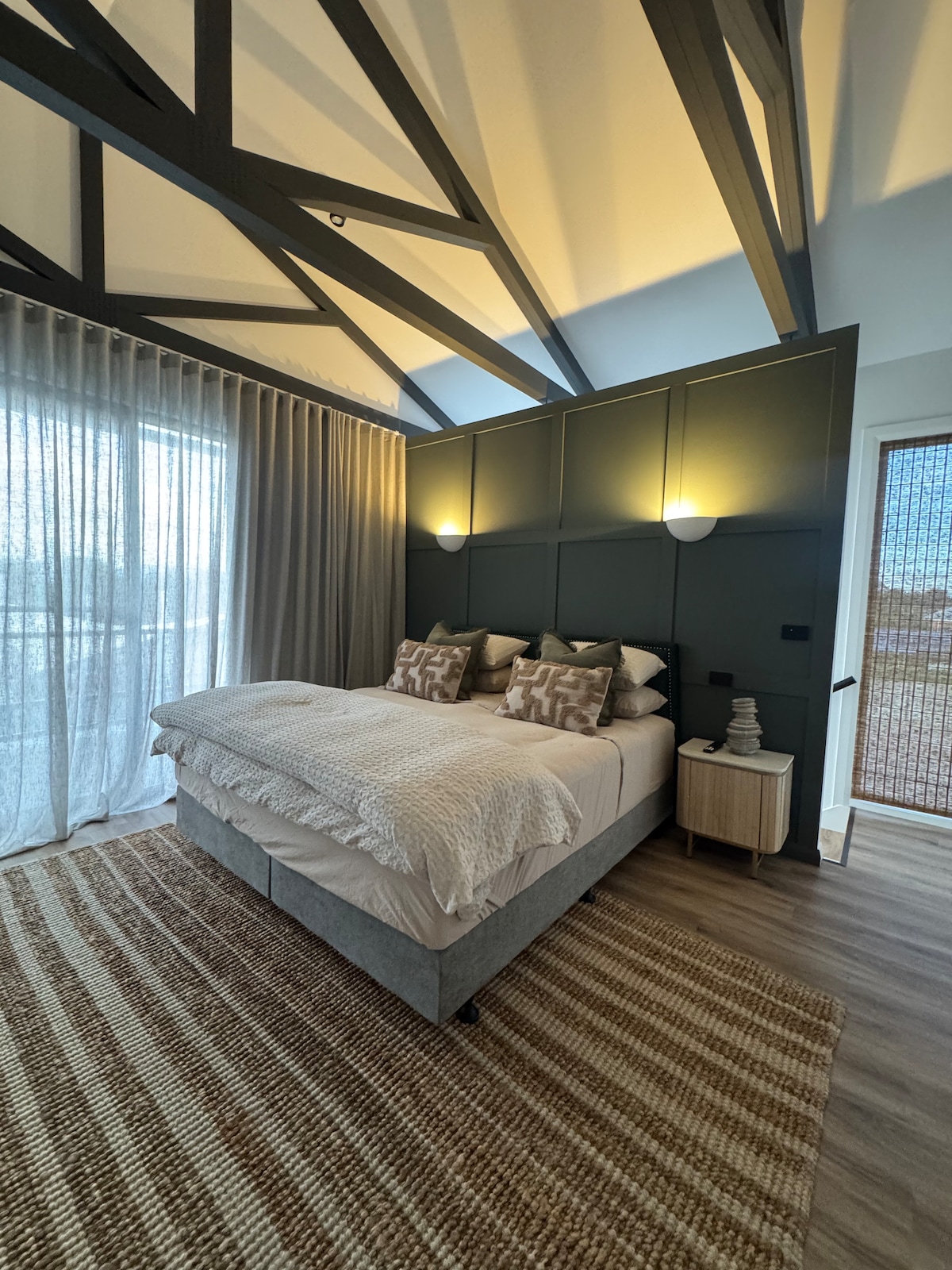The bedroom features a spacious bed adorned with decorative pillows, complemented by warm lighting from the wall sconces. A large window with sheer curtains allows natural light to fill the room, while a textured area rug adds warmth to the wooden floor.