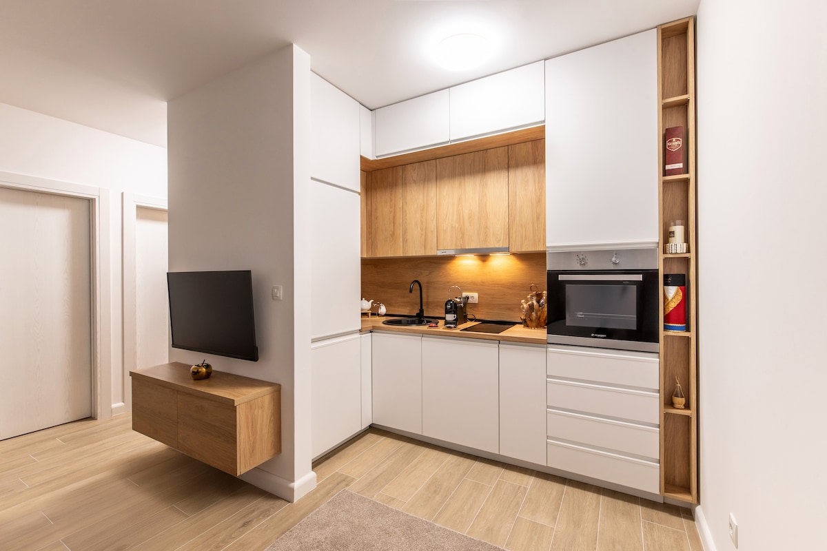 A compact kitchen is highlighted, featuring modern cabinetry in white and light wood. Essential appliances, including a microwave and stovetop, are neatly integrated. A flat-screen TV is mounted on the wall, offering a streamlined appearance. Soft lighting enhances the warm tones of the wood.