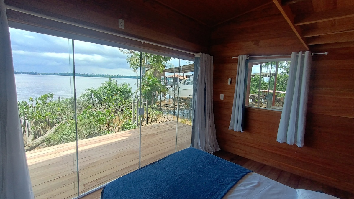 A room with wooden walls features large glass sliding doors that open to a view of a serene river and lush greenery. Soft curtains frame the windows, while a cozy bed is positioned in the foreground, inviting relaxation.