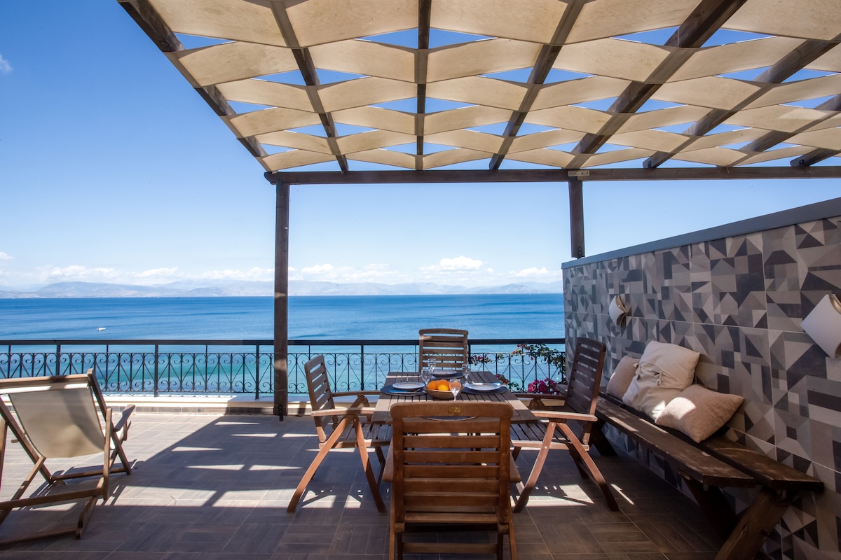 An outdoor seating area is offered under a patterned canopy, featuring a wooden table and multiple lounge chairs. The Ionian Sea is visible in the backdrop, framed by a balcony railing. The sky is clear, with distant mountains seen on the horizon.