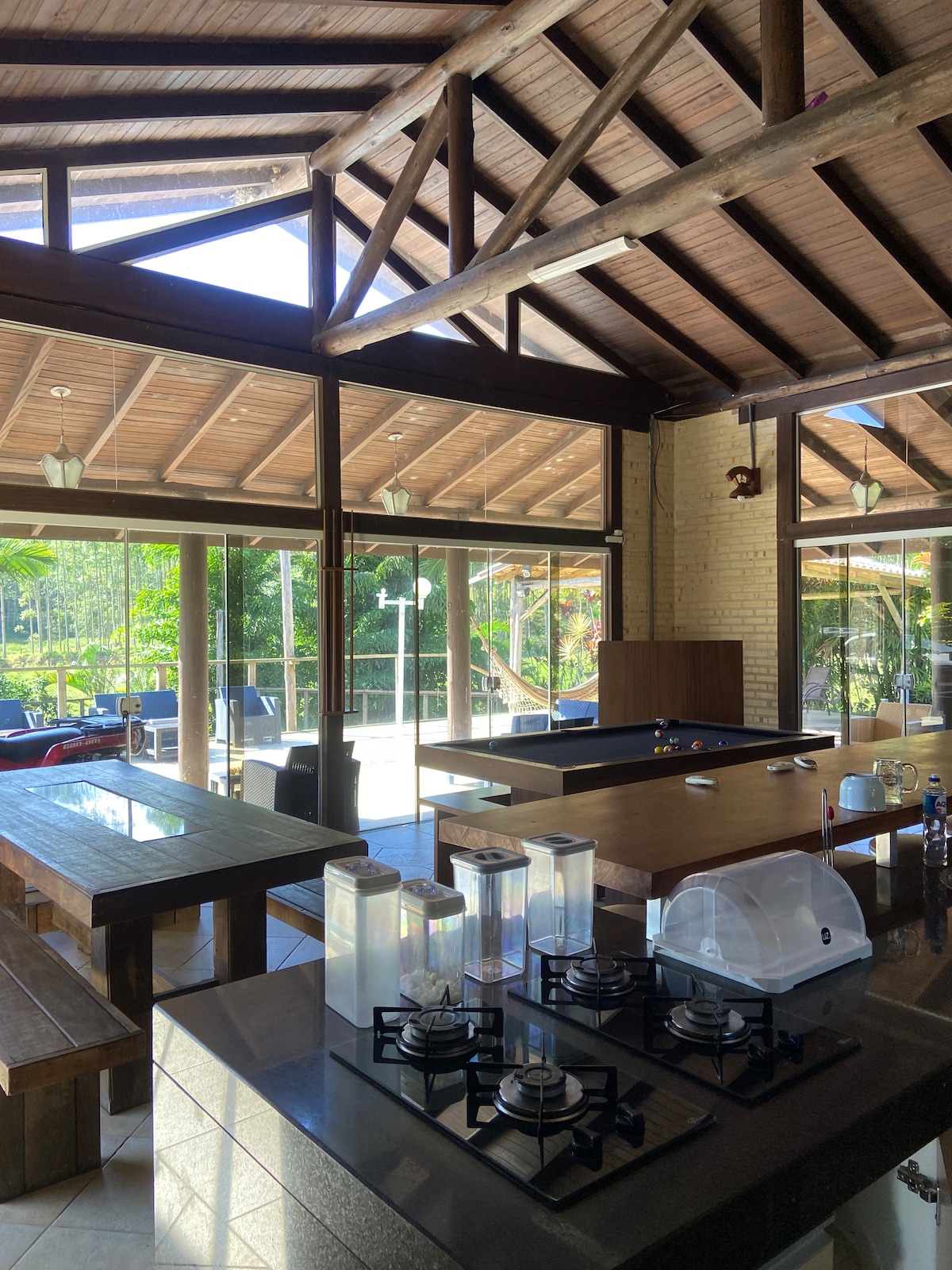 A spacious kitchen area features wooden beams overhead and large windows allowing natural light. A stovetop is positioned in front, while a dining table with ample seating is visible nearby. A billiard table is present, along with views of the lush outdoor surroundings.