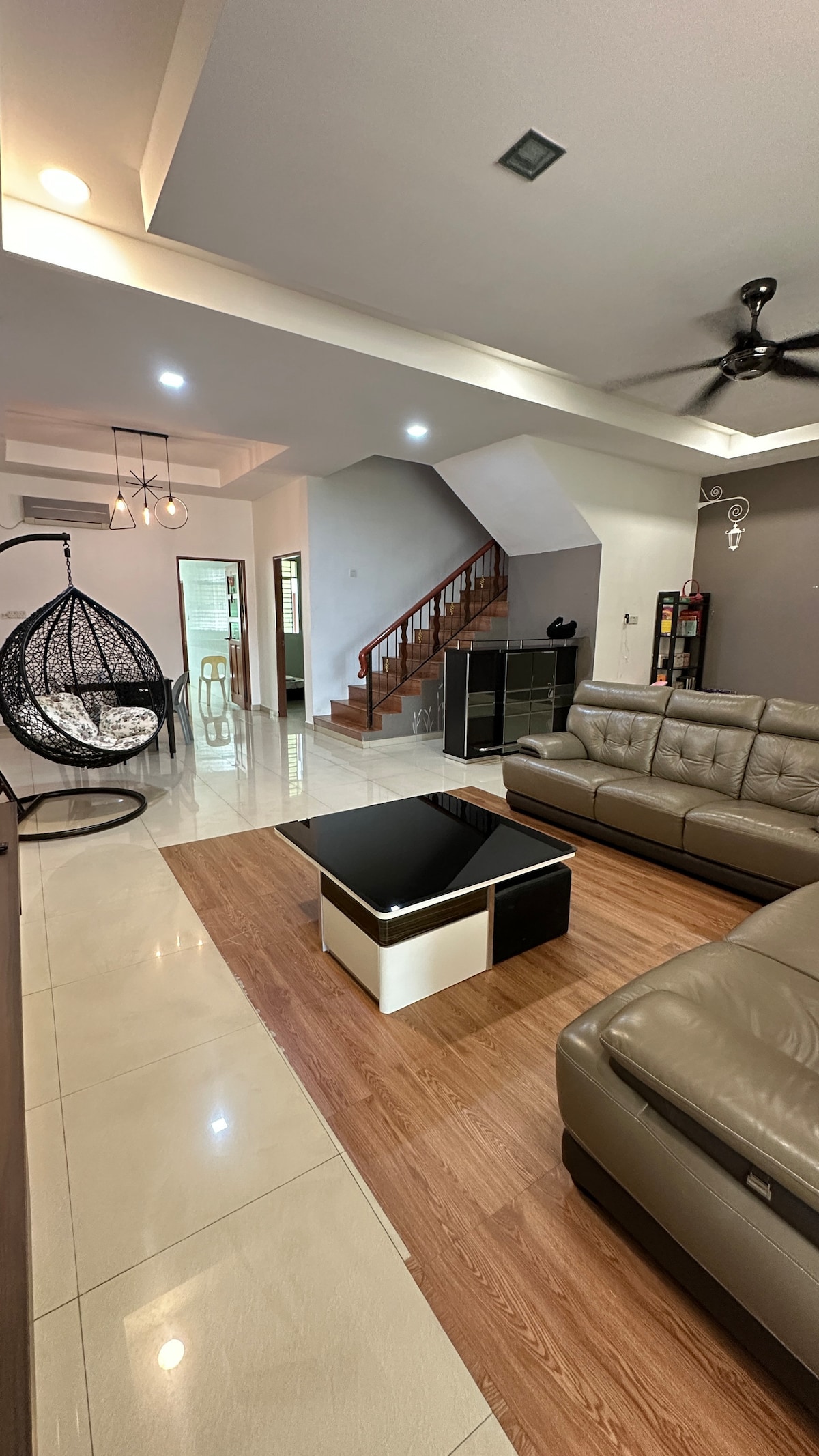 A spacious living area showcases modern furnishings, including a sleek coffee table and comfortable sofa. A stylish hanging chair adds character. The area is brightly lit, with a staircase leading to the upper levels visible in the background.