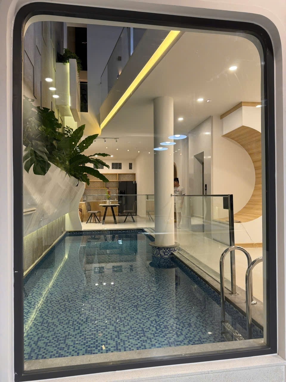 A serene view of a private swimming pool is framed by a large window. Tropical plants are visible along the walls, and a dining area with a table and chairs is revealed in the background, illuminated by modern lighting. The space has a clean, contemporary design.