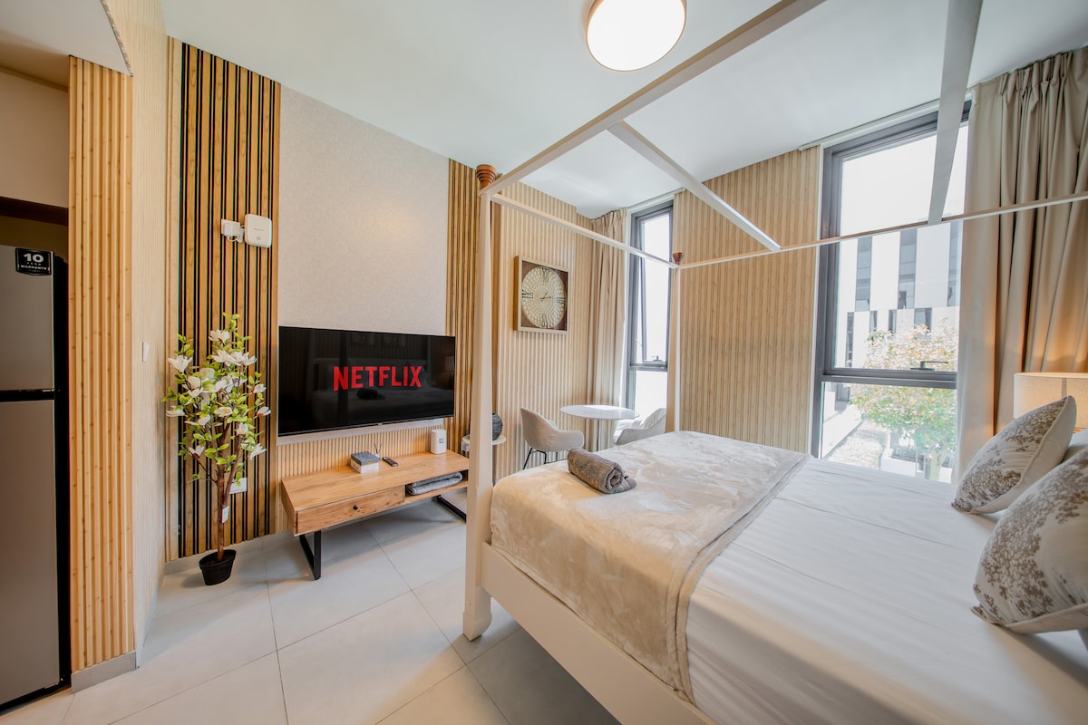 A contemporary bedroom is fitted with a spacious bed and a flat-screen television mounted on a wooden accent wall. A small dining table is positioned near a window, complemented by two chairs. Natural light enters the room, which features minimalist decor and soft neutral tones.