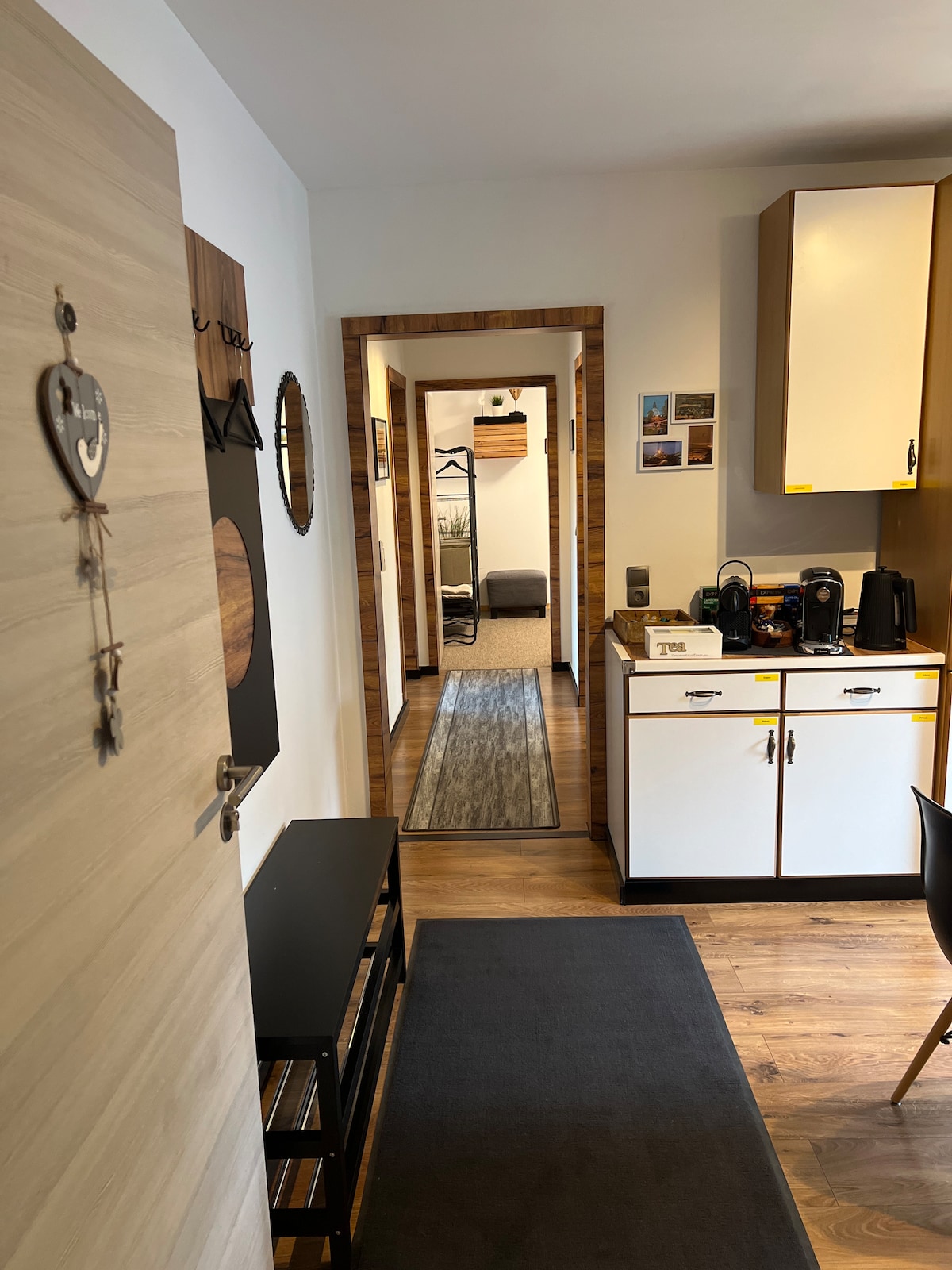 A small hallway connects various areas of the apartment, featuring wooden flooring and a gray rug. On the right, a kitchenette is visible with cabinets and coffee-making facilities. A cozy seating area is seen in the background, enhancing the inviting layout of the space.
