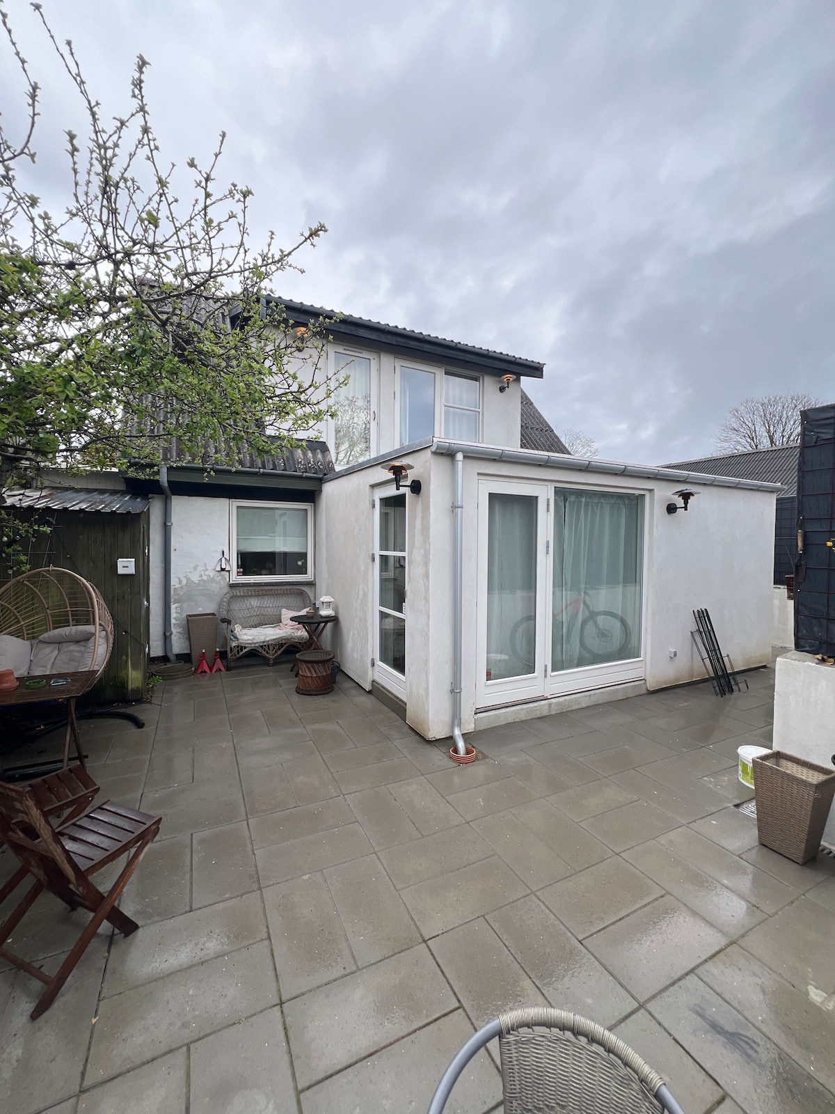 The outdoor patio area is paved with large tiles and features a glass-walled extension at the rear. A seating arrangement with wooden chairs and a table is visible, surrounded by greenery. The cloudy sky adds a subdued light to the space.